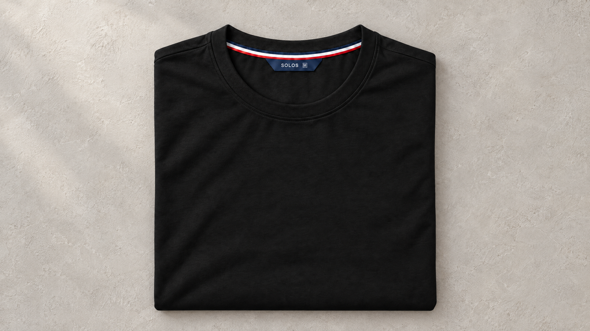 Folded black SOLOS crewneck T-shirt with the approved navy neck label and striped tape on a warm grey surface