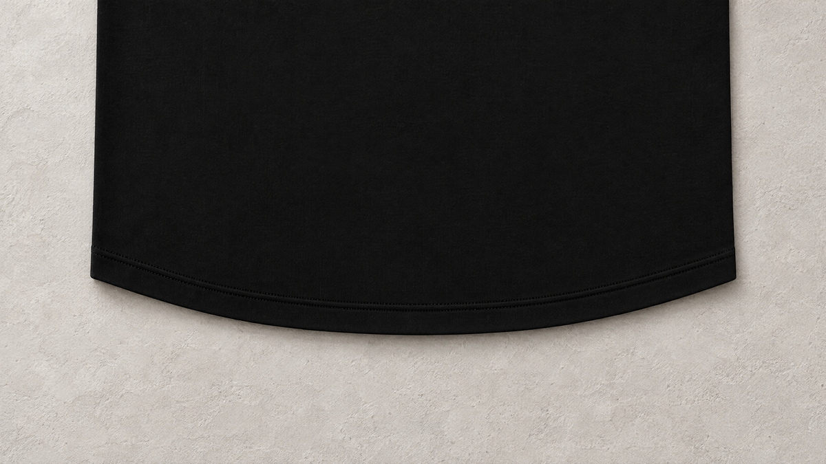Close-up of the subtle curved hem and double stitching of a black SOLOS T-shirt on a warm grey surface
